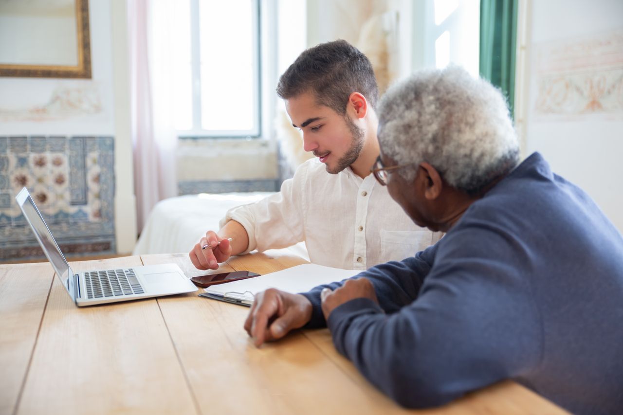 contact-img Young man assisting senior adult with laptop at home for social needs.